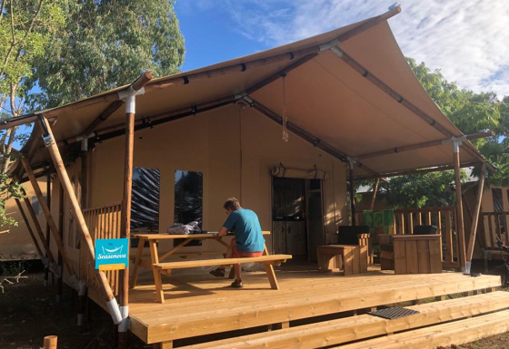 A person sits at a picnic table on a wooden deck outside a safari tent, surrounded by trees and sky.