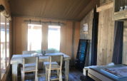 Vista interna di una tenda safari con tavolo da pranzo in legno rustico, sedie e luce soffusa.