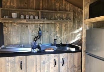 Small kitchen area inside a safari tent featuring wooden walls, cups, glasses, a sink, and a fridge.