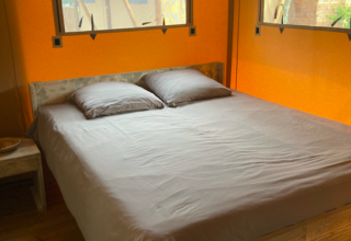Cama cómoda con sábanas grises en el interior de una tienda safari, con piso de madera y paredes naranjas.