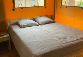 Cama cómoda con sábanas grises en el interior de una tienda safari, con piso de madera y paredes naranjas.