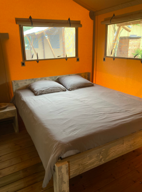 Cama cómoda con sábanas grises en el interior de una tienda safari, con piso de madera y paredes naranjas.