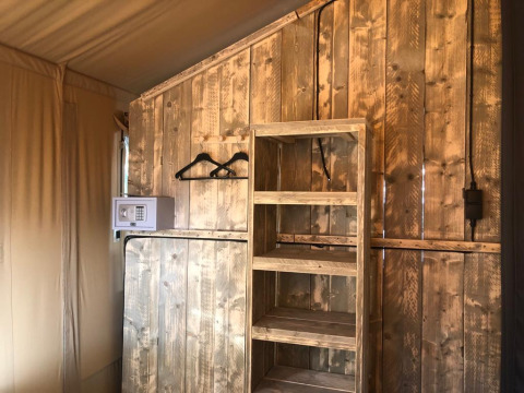 Safari tent interior with wooden shelves, clothes hangers, and safe, set against rustic wooden walls.
