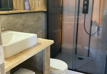 Baño moderno en tienda safari con paredes de madera, ducha de cristal, inodoro y lavabo de sobre madera.