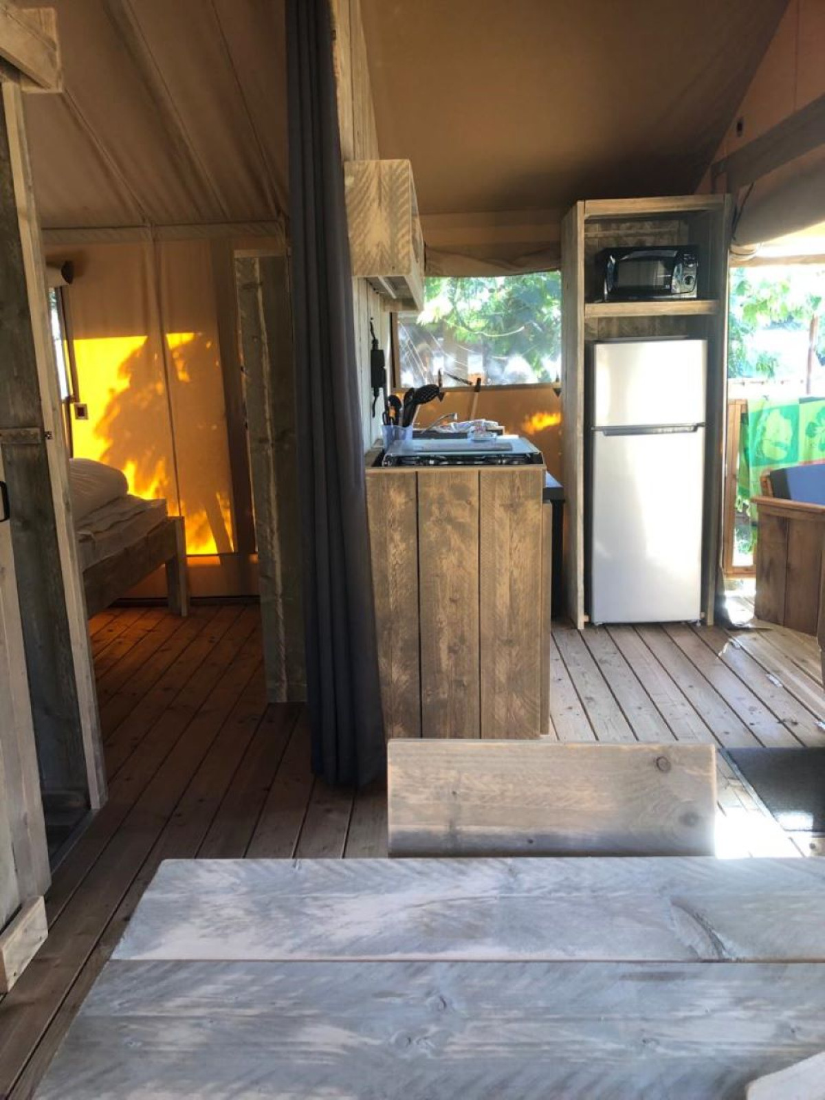 Interieur van een safaritent met houten meubels, kitchenette, koelkast en een zonnige slaapruimte.