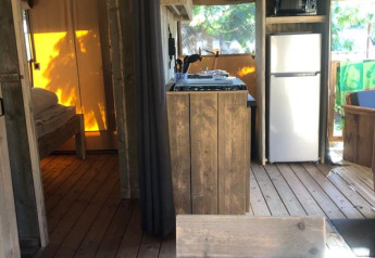Interno di tenda safari con arredamento in legno, angolo cottura, frigorifero e camera illuminata dal sole.
