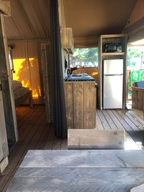 Safari tent interior featuring wooden furniture, a compact kitchen, refrigerator, and sunlit bedroom area.