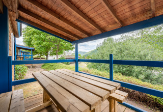 Terraza de madera techada en alojamiento glamping con mesa y bancos, vistas a árboles y colinas verdes.