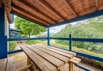 Covered wooden terrace at glamping site with picnic table and benches, overlooking lush greenery.