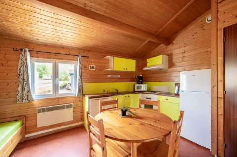 Cozy glamping accommodation kitchen with wooden interior, round table, fridge, window and yellow cabinets.