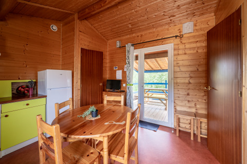 Interior de una acogedora cabaña glamping con paredes de madera, mesa redonda, nevera y terraza exterior.