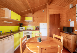 Cocina y comedor glamping con paneles de madera, armarios amarillos, nevera, mesa y televisor.