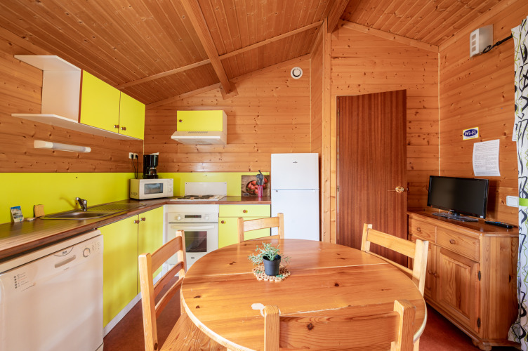 Cocina y comedor glamping con paneles de madera, armarios amarillos, nevera, mesa y televisor.