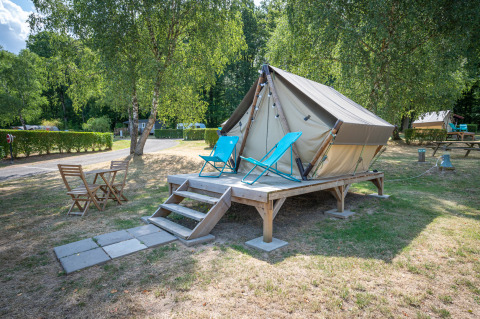 Photo d'une tente tipi sur une terrasse en bois avec chaises longues et table sur un terrain de camping verdoyant.