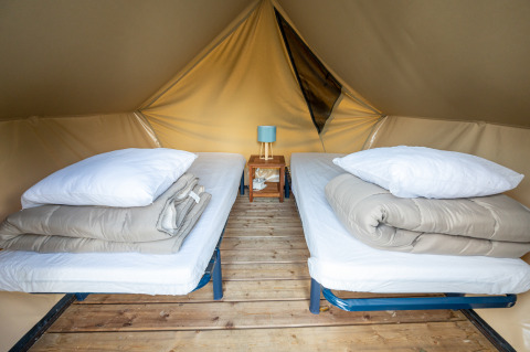 Inside view of a teepee tent with two single beds, wooden floor, and small table with a lamp between them.