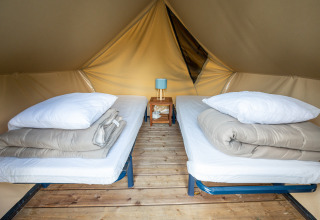 Indersiden af et tipi-telt med to enkeltsenge, trægulv og et lille bord med lampe og kop i midten.