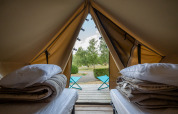 Indersiden af et tipi-telt med to senge, puder, tæpper og udsigt til terrasse og natur udenfor.