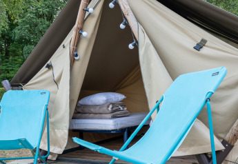 Tienda tipo tipi con ropa de cama y dos sillas azules sobre una plataforma de madera al aire libre.