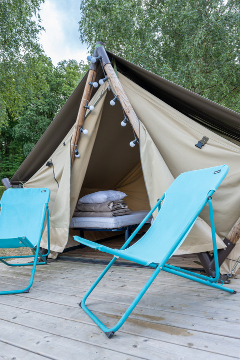 Tipi-style tent with cozy bedding and two blue chairs on a wooden deck surrounded by trees outside.