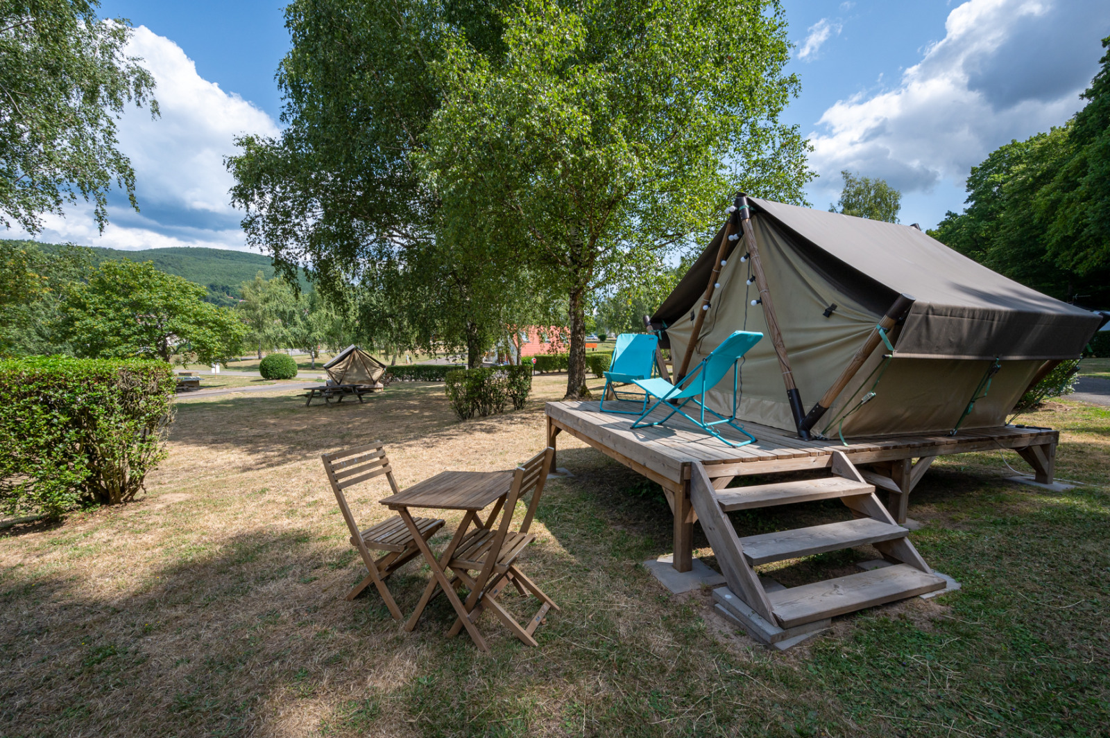 Een tipi-tent op een houten platform met stoelen en tafel, omringd door bomen en grasveld.