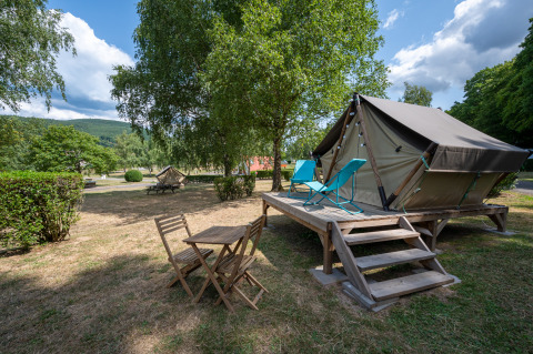 Tienda tipi sobre plataforma de madera con sillas y mesa, rodeada de árboles y césped bajo cielo nublado.