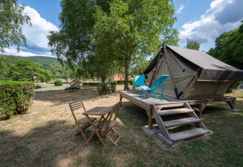 Tienda tipi sobre plataforma de madera con sillas y mesa, rodeada de árboles y césped bajo cielo nublado.