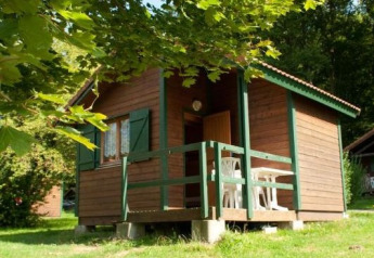 Houten chaletje met groene afwerking en veranda, liggend in een groene bosrijke omgeving met zon.