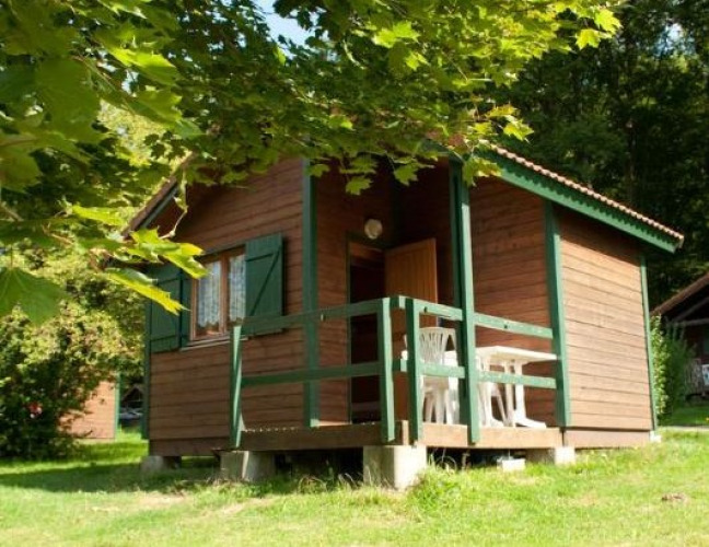 Cabaña de madera con terraza y sillas blancas de plástico, rodeada de árboles verdes y naturaleza.