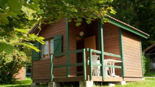 Cabaña de madera con terraza y sillas blancas de plástico, rodeada de árboles verdes y naturaleza.