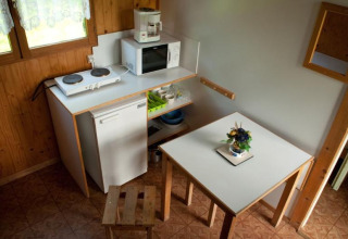 Pequeña cocina en una cabaña con mesa, taburetes, placa eléctrica, nevera, microondas y escurreplatos.