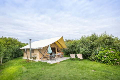 Safarizelt namens Tente Trappeur auf einer grünen Wiese, umgeben von Büschen, unter bewölktem Himmel.