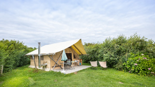 Tienda safari llamada Tente Trappeur sobre césped, rodeada de arbustos y bajo un cielo nublado.