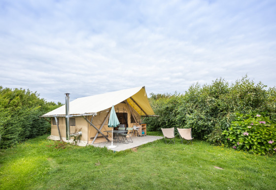 Safarizelt namens Tente Trappeur auf einer grünen Wiese, umgeben von Büschen, unter bewölktem Himmel.