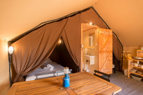 Interno di una tenda safari con letto, tavolo in legno, bagno privato e tende marroni aperte.