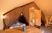 Interno di una tenda safari con letto, tavolo in legno, bagno privato e tende marroni aperte.