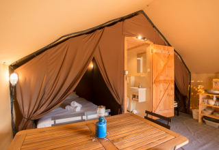 Interior of a safari tent with a bed, wooden table, private bathroom, and open brown tent curtains.