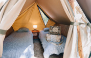 Interior de una tienda tipi con dos camas individuales, una mesita con lámpara y una maleta decorativa.