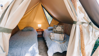 Interior de una tienda tipi con dos camas individuales, una mesita con lámpara y una maleta decorativa.