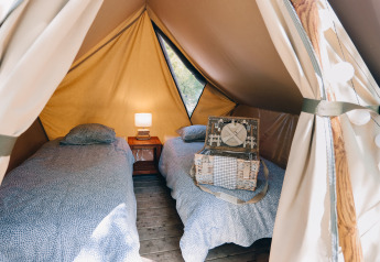 Interior de una tienda tipi con dos camas individuales, una mesita con lámpara y una maleta decorativa.
