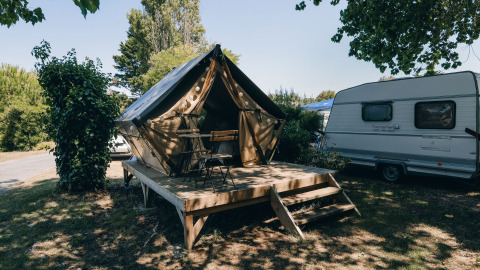 Een foto van een tipi-tent met houten terras en stoelen, naast een caravan op een schaduwrijke camping.