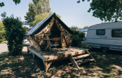 Foto de una tienda tipi con terraza de madera y sillas, junto a una caravana en un camping arbolado.