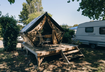Foto van een tipi-tent met houten terras en stoelen, naast een caravan op een groene kampeerplaats.