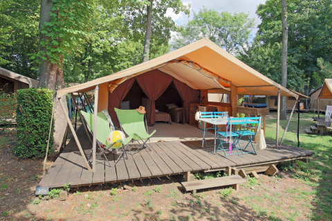 Safari tent Bungalow Classic featuring wooden deck, chairs, and table on a lush, shaded campground setting.