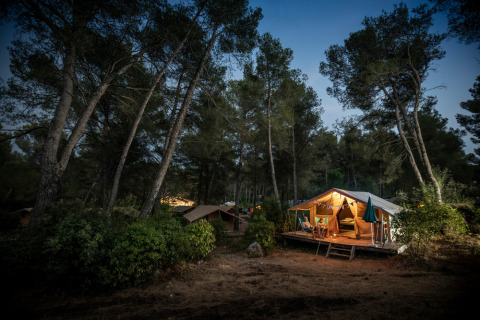 Tienda safari Bungalow tent Classic al anochecer en el bosque, iluminada y gente relajada en la terraza.