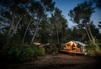 Tienda safari Bungalow tent Classic al anochecer en el bosque, iluminada y gente relajada en la terraza.