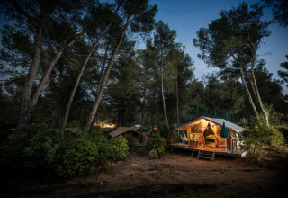 Safari-Bungalowzelt Classic im Wald am Abend, beleuchteter Eingang, Menschen sitzen auf der Veranda.