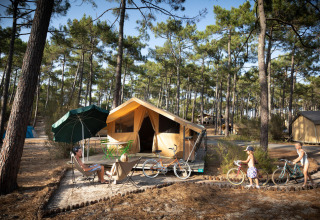 Familia relajándose en un Bungalow tent Classic en el bosque, niños jugando y bicicletas cerca de la tienda.