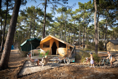 Famille se détend devant une tente safari Bungalow Classic en forêt, enfants et vélos à proximité du camp.