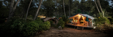 Tienda tipo bungalow Classic iluminada por la noche en Huttopia Landes Sud, Francia, personas relajándose frente a la tienda.