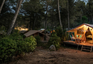 Tienda tipo bungalow Classic iluminada por la noche en Huttopia Landes Sud, Francia, personas relajándose frente a la tienda.